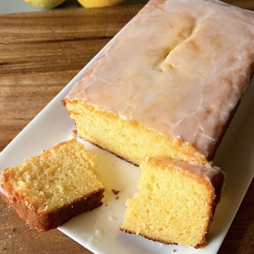 A delicious lemon loaf cake in a plate with a slice broken in half. The cake is yellow in colour.