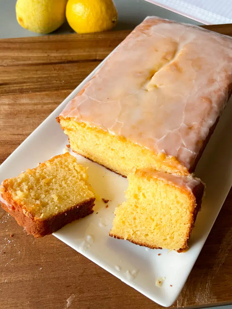 A delicious lemon loaf cake in a plate with a slice broken in half. The cake is yellow in colour.