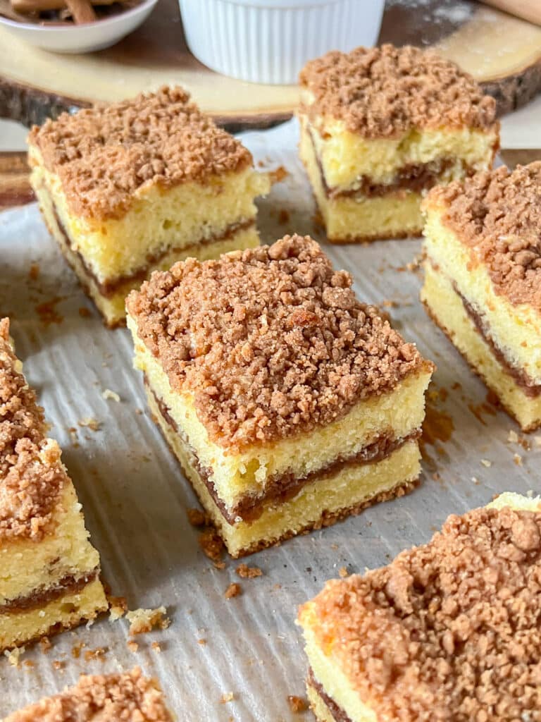 A slice of cinnamon coffee cake on an off white surface, surrounded by more slices of cake