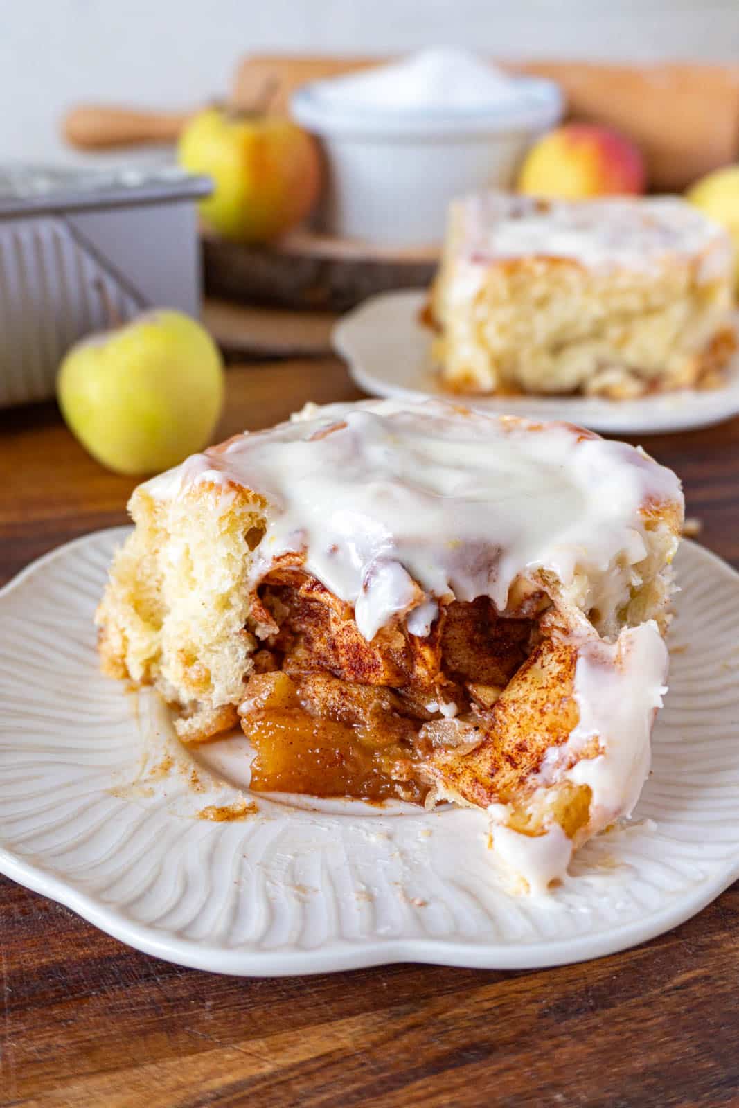 a torn-into cinnamon roll on w white plate with apples in the background