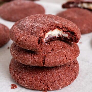 3 stacked stuffed red velvet cookies on a white surface with a couple more in the back. The top one has a bite taken out of it