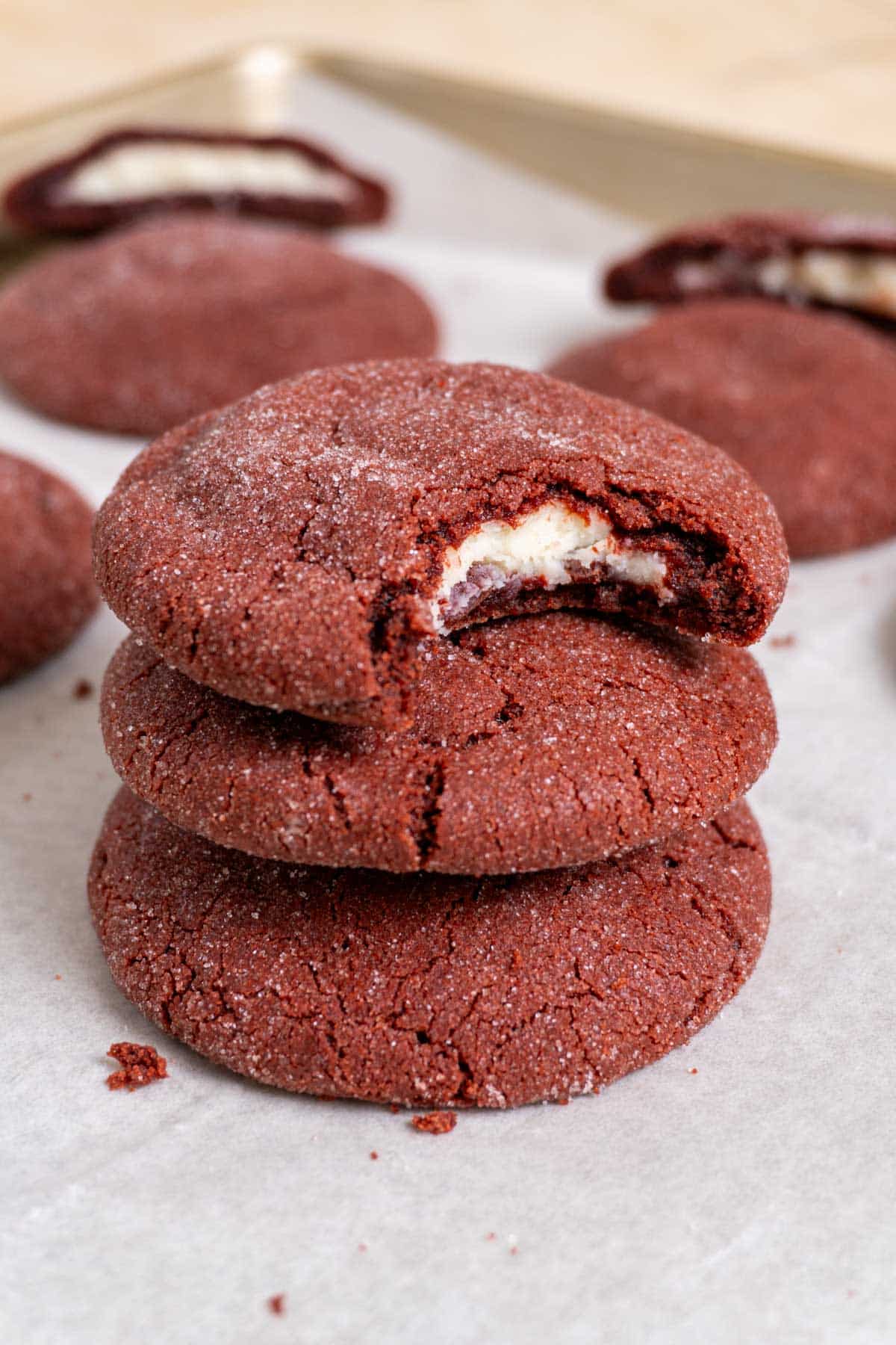 3 stacked stuffed red velvet cookies on a white surface with a couple more in the back. The top one has a bite taken out of it