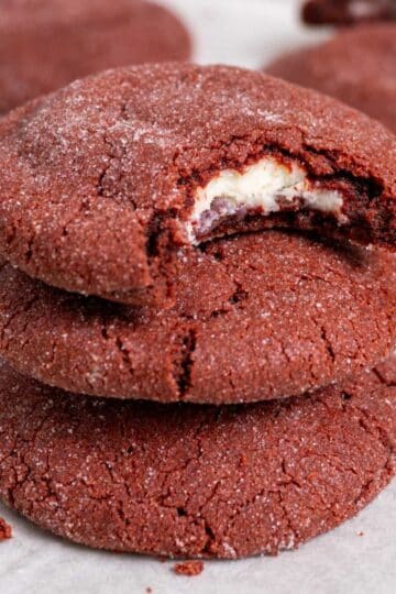 3 stacked stuffed red velvet cookies on a white surface with a couple more in the back. The top one has a bite taken out of it