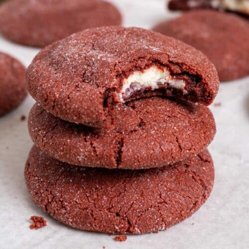 3 stacked stuffed red velvet cookies on a white surface with a couple more in the back. The top one has a bite taken out of it