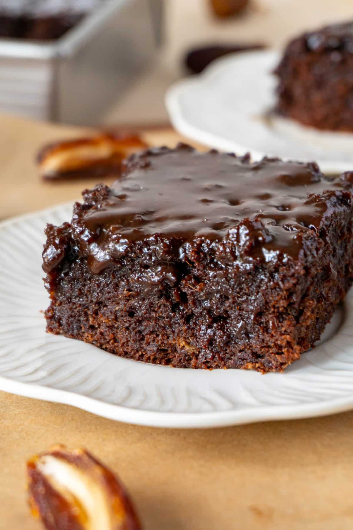 a slice of chocolate sticky toffee cake on a white plate, on a brown surface. Another white plate with a slice is in the background