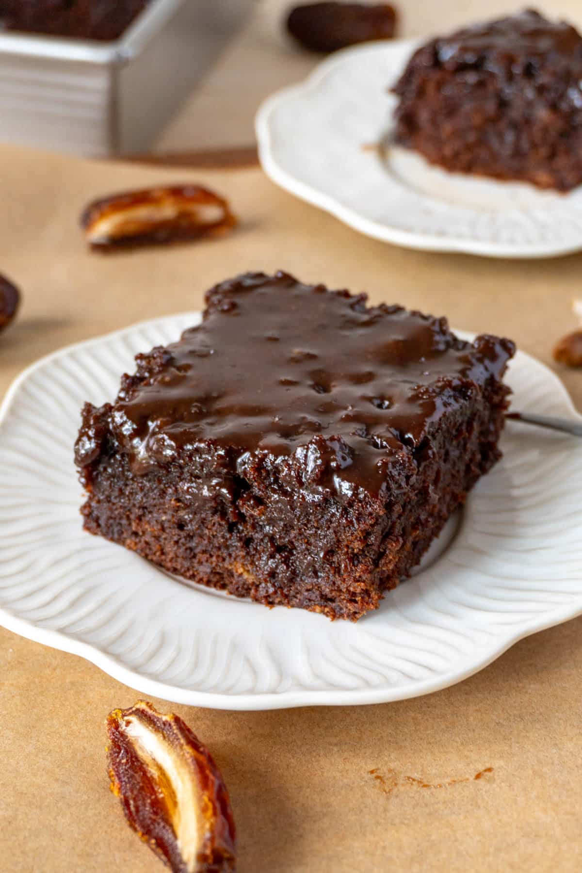 a slice of chocolate sticky toffee cake on a white plate, on a brown surface. Another white plate with a slice is in the background