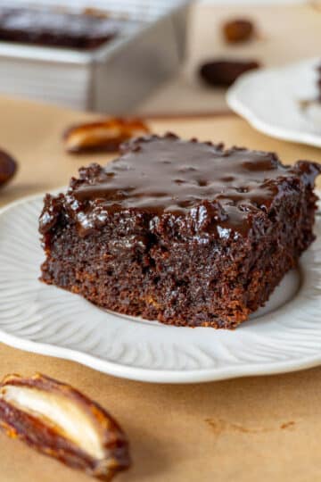 a slice of chocolate sticky toffee cake on a white plate, on a brown surface. Another white plate with a slice is in the background