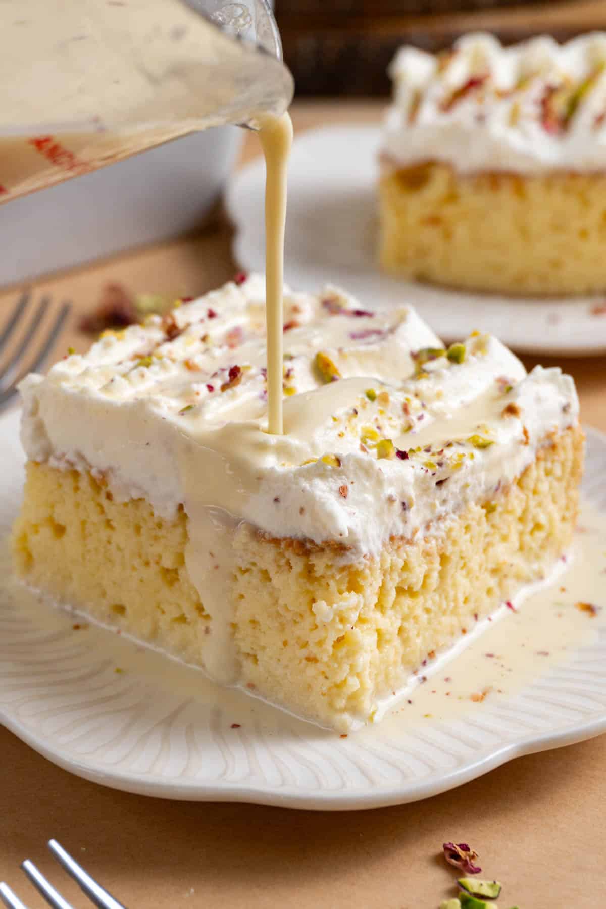 a slice of rasmalai tres leches on a white plate. There's the milk mixture being poured onto the cake.